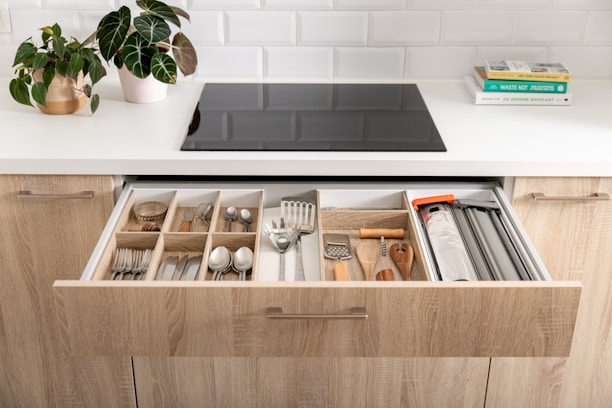 A modern kitchen scene featuring an open drawer filled with organized kitchen utensils and cutlery, including forks, spoons, knives, and various cooking tools. The countertop above the drawer holds a potted plant and a couple of stacked books on one side, with a sleek black stove embedded in the white countertop. The kitchen cabinets are made of light wood and there's a white tiled backsplash creating a clean and contemporary look.