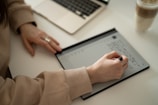 A person is writing on an electronic tablet with a stylus, with a laptop nearby on a desk. They are wearing a beige sweater and a ring, and there's a cup with a drink to the side.