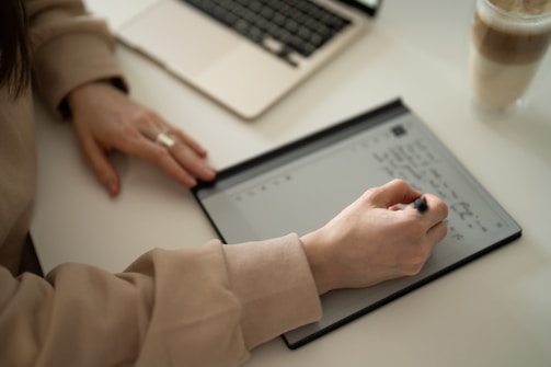 A person is writing on an electronic tablet with a stylus, with a laptop nearby on a desk. They are wearing a beige sweater and a ring, and there's a cup with a drink to the side.