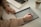 A person is writing on an electronic tablet with a stylus, with a laptop nearby on a desk. They are wearing a beige sweater and a ring, and there's a cup with a drink to the side.