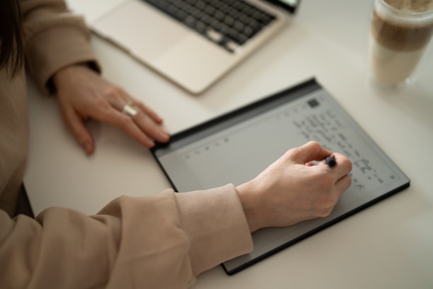 A person is writing on an electronic tablet with a stylus, with a laptop nearby on a desk. They are wearing a beige sweater and a ring, and there's a cup with a drink to the side.