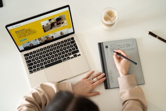 a person is writing on a notebook while using a laptop
