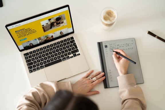 A person is writing notes on a digital tablet with a stylus, while a laptop displaying a webpage is open nearby. A glass of coffee sits on the desk, and a pen lies next to it. The setting suggests a workspace or study environment with a focus on multitasking and digital tools.