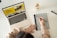 A person is writing notes on a digital tablet with a stylus, while a laptop displaying a webpage is open nearby. A glass of coffee sits on the desk, and a pen lies next to it. The setting suggests a workspace or study environment with a focus on multitasking and digital tools.