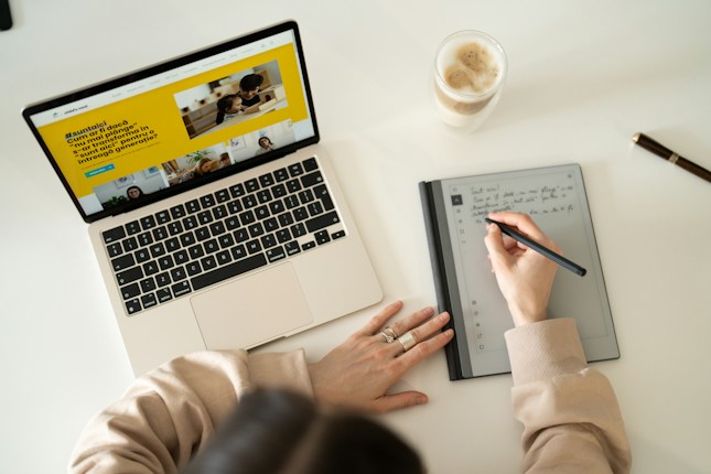 A person is writing notes on a digital tablet with a stylus, while a laptop displaying a webpage is open nearby. A glass of coffee sits on the desk, and a pen lies next to it. The setting suggests a workspace or study environment with a focus on multitasking and digital tools.
