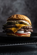 A juicy double cheeseburger is placed on a dark textured surface. It contains two beef patties layered with melted cheese, slices of pickle, fresh onions, and is topped with a soft, golden bun. The lighting highlights the textures and details of the ingredients.