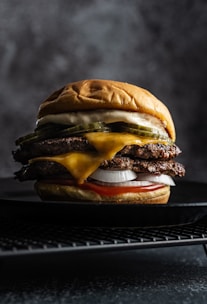 A juicy double cheeseburger is placed on a dark textured surface. It contains two beef patties layered with melted cheese, slices of pickle, fresh onions, and is topped with a soft, golden bun. The lighting highlights the textures and details of the ingredients.