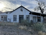An old, single-story building with a gabled roof stands in a rural setting. The exterior wall appears worn and weathered, with peeling white paint. There are several windows with aging frames, and a chain-link fence running in front of the building. A sign on the fence reads 'No Trespassing.' Overgrown shrubs and dry grass cover the foreground. A leafless tree is visible to the right side of the image.