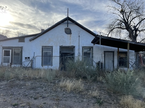 An old, single-story building with a gabled roof stands in a rural setting. The exterior wall appears worn and weathered, with peeling white paint. There are several windows with aging frames, and a chain-link fence running in front of the building. A sign on the fence reads 'No Trespassing.' Overgrown shrubs and dry grass cover the foreground. A leafless tree is visible to the right side of the image.