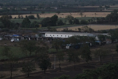 A rural landscape with fields and sparse trees in the foreground. A long, white building and several smaller structures are visible, surrounded by greenery and farmland.