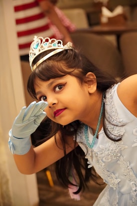 A young girl dressed in a blue, lace-embroidered dress with matching gloves and a jeweled tiara is posing playfully. She is glancing sideways with her hand raised near her forehead in a gesture of curiosity or salutation.