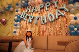 A young girl dressed in a princess gown and tiara sits on a couch beneath large, metallic blue balloons spelling out 'HAPPY BIRTHDAY'. Surrounding her are decorations consisting of blue and white balloons and metallic gold streamers.