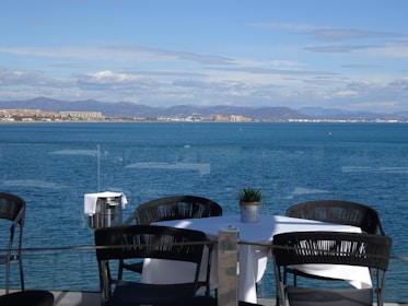 A table set with a white cloth and a small potted plant is positioned on an outdoor patio overlooking a vast, calm sea. Several dark wicker chairs surround the table. In the distance, there is a view of a coastline with buildings and mountains under a partly cloudy sky. A metal ice bucket with a white cloth draped over it is nearby.