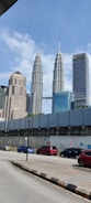 Tall skyscrapers dominate the skyline with the Petronas Towers prominently featured. Below, a street is lined with colorful cars and a barricaded area with flags and murals. A clear, sunny sky provides a backdrop.