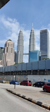 Tall skyscrapers dominate the skyline with the Petronas Towers prominently featured. Below, a street is lined with colorful cars and a barricaded area with flags and murals. A clear, sunny sky provides a backdrop.