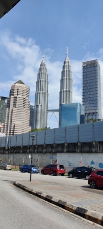 Tall skyscrapers dominate the skyline with the Petronas Towers prominently featured. Below, a street is lined with colorful cars and a barricaded area with flags and murals. A clear, sunny sky provides a backdrop.