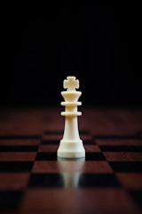 A white chess king piece stands prominently on a checkered wooden chessboard with a dark background, casting a reflection on the surface.