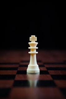 A white chess king piece stands prominently on a checkered wooden chessboard with a dark background, casting a reflection on the surface.