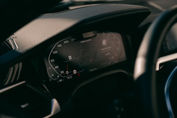 A close-up view of a car's digital dashboard display, showing various informational panels including a speedometer and GPS navigation system. The dashboard is modern with illuminated numbers and sleek design.