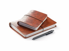 Two brown leather notebooks with intricate topographic designs embossed on the covers are stacked together. A black pen lies next to the notebooks.
