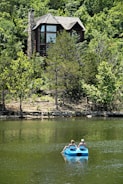A wooden house with large windows sits nestled among lush green trees. In the foreground, two people are leisurely paddling on a blue paddle boat in a calm lake.