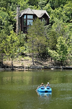 A wooden house with large windows sits nestled among lush green trees. In the foreground, two people are leisurely paddling on a blue paddle boat in a calm lake.