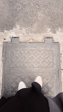A person stands on a patterned metal cover with 'KOTA SURAKARTA' inscribed on it, set against a textured grey and sandy surface.