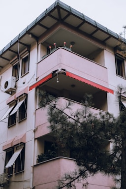 A multi-story residential building with balconies on each floor, partially obscured by pine tree branches. It has a light pink exterior with red trim, featuring an air conditioning unit attached to one side. Potted plants are visible on some of the balconies.