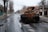 A street scene with a heavily damaged, rusted, and destroyed armored vehicle. The setting is a desolate urban area with leafless trees lining the street. The road is muddy and wet, suggesting recent rainfall. In the distance, there are other vehicles and damaged structures, indicating possible conflict or abandonment.