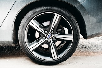 A close-up view of a car wheel featuring a five-spoke alloy rim branded with a logo at the center. The tire has visible tread and branding along the sidewall, while the car's body is slightly dusty. The background includes a sandy surface.