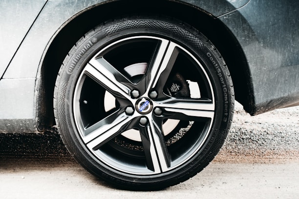 A close-up view of a car wheel featuring a five-spoke alloy rim branded with a logo at the center. The tire has visible tread and branding along the sidewall, while the car's body is slightly dusty. The background includes a sandy surface.