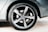 A close-up view of a car wheel featuring a five-spoke alloy rim branded with a logo at the center. The tire has visible tread and branding along the sidewall, while the car's body is slightly dusty. The background includes a sandy surface.