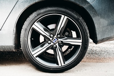 A close-up view of a car wheel featuring a five-spoke alloy rim branded with a logo at the center. The tire has visible tread and branding along the sidewall, while the car's body is slightly dusty. The background includes a sandy surface.