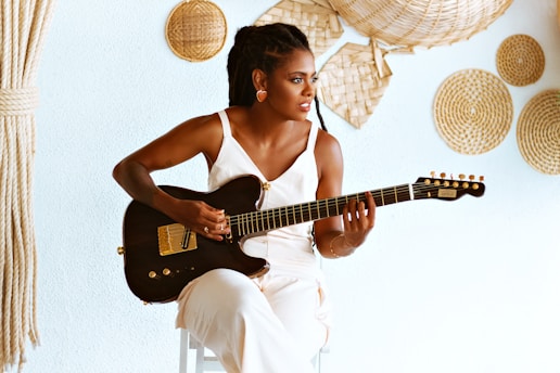 A woman is seated and playing a black electric guitar. She is wearing a white outfit and is surrounded by decorative straw circles on a light blue wall. Her hair is styled in braids, and the setting has a natural, bohemian feel.