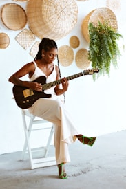 A woman with long braided hair is playing an electric guitar while seated on a white chair. She is wearing a white sleeveless outfit and green strappy sandals. Behind her, there is a white wall decorated with various woven baskets and a hanging green plant.