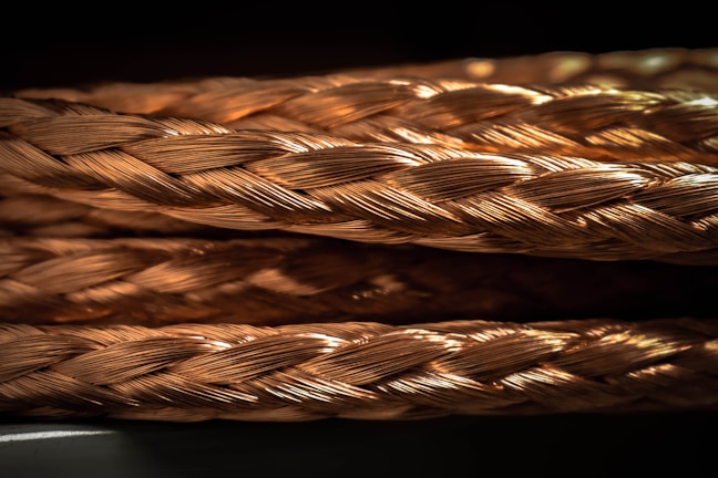 Close-up view of intricately braided copper wires with a shiny, metallic finish. The weaving pattern is detailed and reflects light, creating a warm, golden appearance.