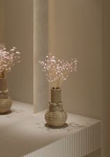 A decorative vase with a textured geometric design is placed on a minimalist surface. The vase contains delicate, light-colored flowers with small blossoms. The lighting is soft and casts gentle shadows on the vase and surface, creating a serene and elegant atmosphere.