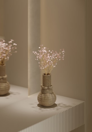 A decorative vase with a textured geometric design is placed on a minimalist surface. The vase contains delicate, light-colored flowers with small blossoms. The lighting is soft and casts gentle shadows on the vase and surface, creating a serene and elegant atmosphere.