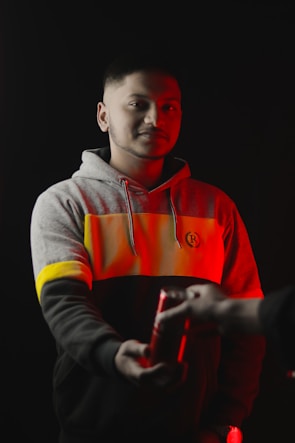 A person wearing a hoodie is handing over a red can, set against a dark background. The image is illuminated with dramatic lighting, highlighting the person's face and the can in shades of red.