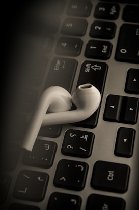 White earbuds are resting on a laptop keyboard, which has a mix of English and Arabic letters. The lighting is dim, creating a focused and moody ambiance.