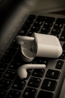 Wireless earbuds rest on a black computer keyboard, with the charging case nearby. The focus is on the earbuds and case, highlighting their sleek white design against the contrasting dark background.