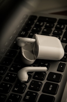 Wireless earbuds rest on a black computer keyboard, with the charging case nearby. The focus is on the earbuds and case, highlighting their sleek white design against the contrasting dark background.
