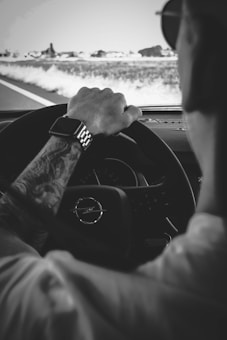 A black and white image of a person driving a car, focusing on their hand on the steering wheel. The driver's arm shows a tattoo and they are wearing a watch. The road ahead is visible through the windshield, with fields on either side.