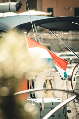 A sailboat with a visible steering wheel and a life preserver attached. A tricolor flag with red, white, and blue stripes is draped on the boat. The background shows a dock or harbor area with blurred elements, possibly including another building or boats. The lighting is warm and sunny, casting soft shadows.