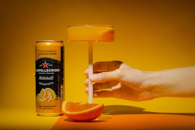 A hand holding a glass filled with an orange-colored drink is positioned beside a can of Sanpellegrino Aranciata on an orange surface. A slice of orange is placed beneath the glass, casting a shadow. The background is a gradient of yellow and orange tones.