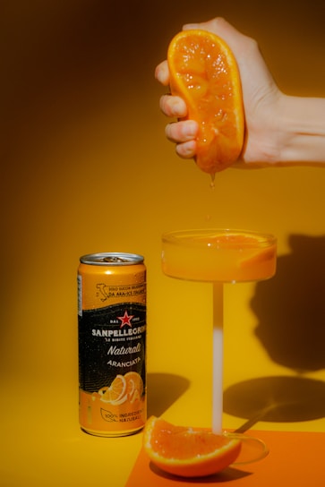A hand is squeezing a fresh orange slice over a glass filled with orange juice. Next to the glass, there is a can of Sanpellegrino Aranciata on an orange surface. The background is in a warm yellow shade, creating a vibrant and refreshing atmosphere.