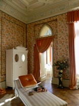 An ornate room with floral-patterned wallpaper and draped curtains in a warm, earthy tone. A chaise longue with a plush orange pillow and a bouquet of pink roses rests on it. A tall white wardrobe with mirrored doors stands against the wall. A vase filled with flowers is nearby, enhancing the elegant and vintage atmosphere of the space.