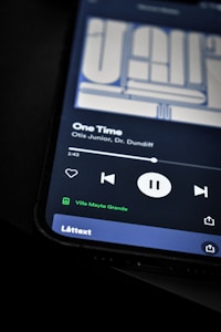 A smartphone screen displaying a music player interface with a song titled 'One Time' by Otis Junior and Dr. Dundiff. The playback controls indicate the song is paused, and text shows it is connected to a device called 'Villa Mayte Grande.' The background is dark, highlighting the screen contents.