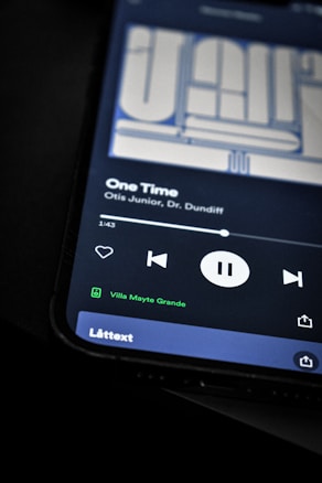 A smartphone screen displaying a music player interface with a song titled 'One Time' by Otis Junior and Dr. Dundiff. The playback controls indicate the song is paused, and text shows it is connected to a device called 'Villa Mayte Grande.' The background is dark, highlighting the screen contents.