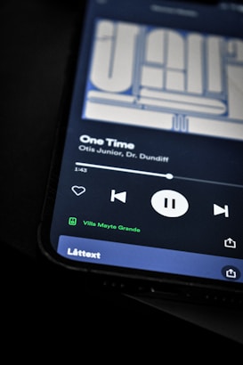 A smartphone screen displaying a music player interface with a song titled 'One Time' by Otis Junior and Dr. Dundiff. The playback controls indicate the song is paused, and text shows it is connected to a device called 'Villa Mayte Grande.' The background is dark, highlighting the screen contents.